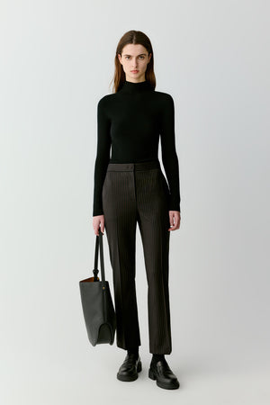 Full-length front view of a model wearing a black ribbed pullover knitted from natural superfine wool. The sweater features a slim silhouette with a mock-neck collar and long sleeves. The ribbed knit structure runs vertically across the garment, creating subtle texture while maintaining softness.