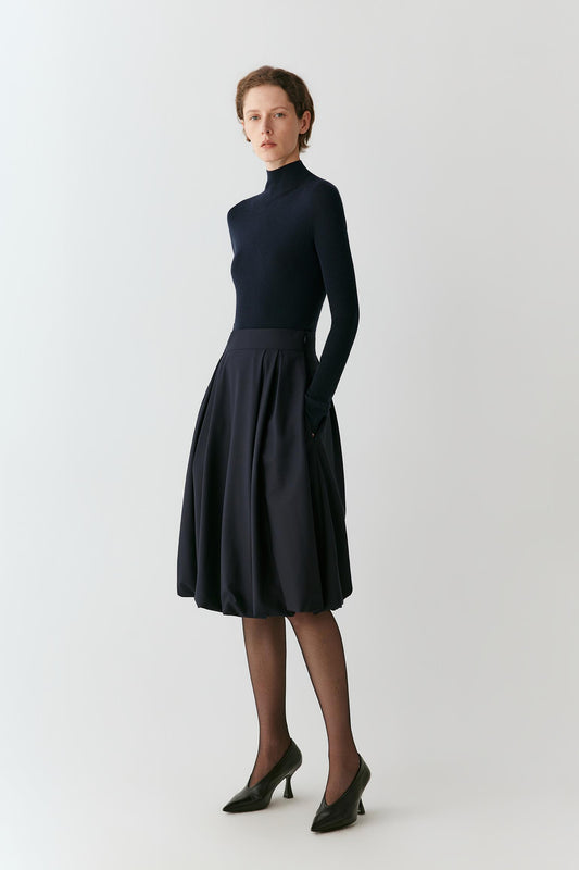 A woman modeling a dark navy blue two-piece outfit consisting of a mock-neck long-sleeve knit top and a matching pleated A-line skirt, paired with black tights and black pointed-toe heels, against a plain light gray studio background.