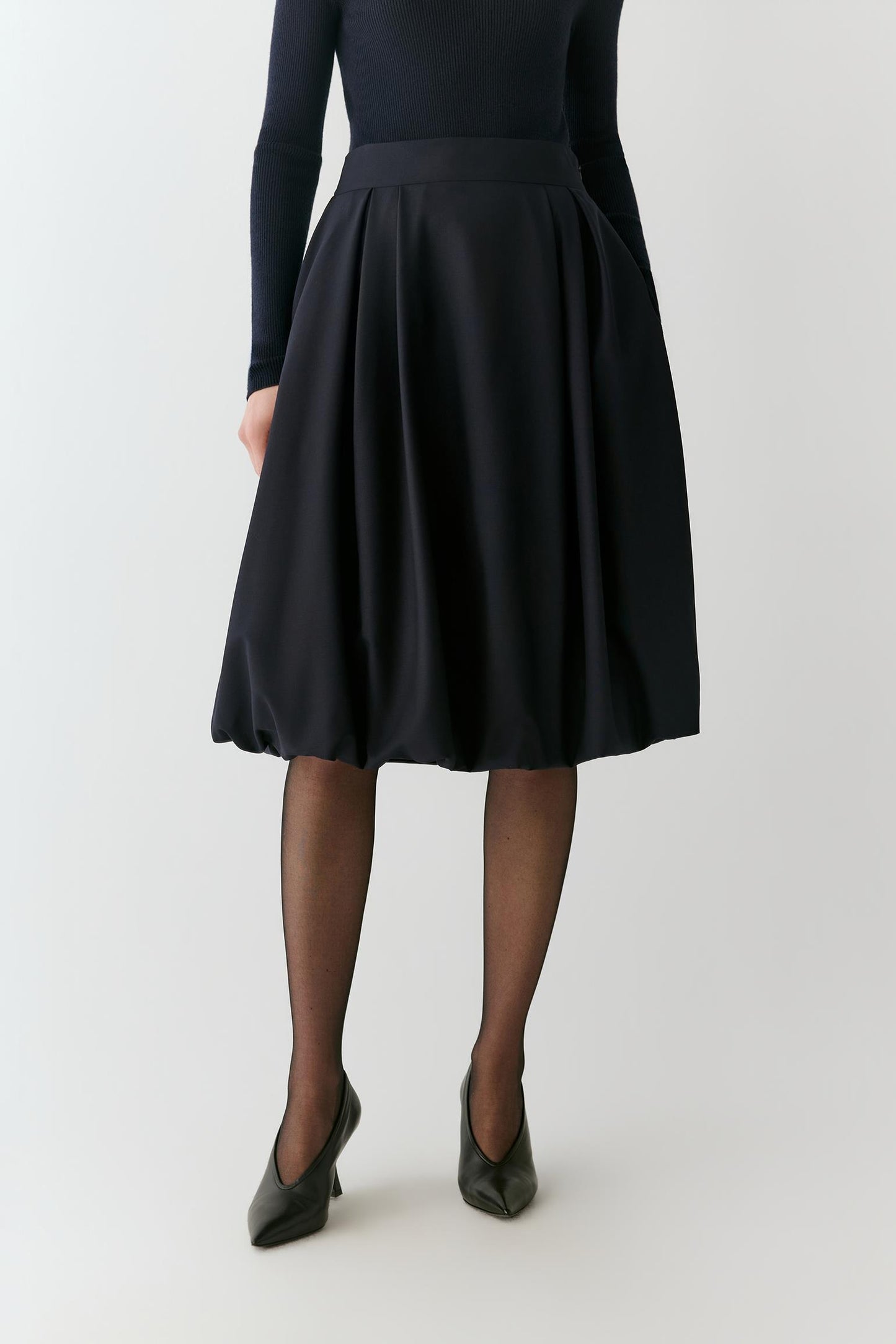 A model is shown from the back wearing a black long-sleeve top, a black pleated A-line midi skirt, sheer black tights, and black pointed-toe heels against a light gray studio background.