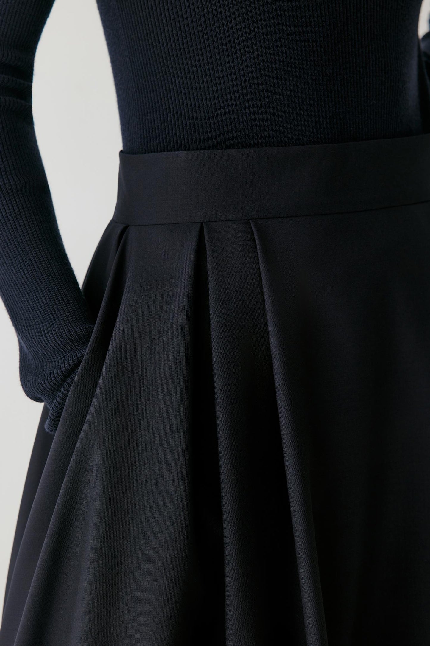 Close-up of a black pleated skirt with a wide waistband, paired with a ribbed black long-sleeve top, shown on a mannequin against a light gray background.