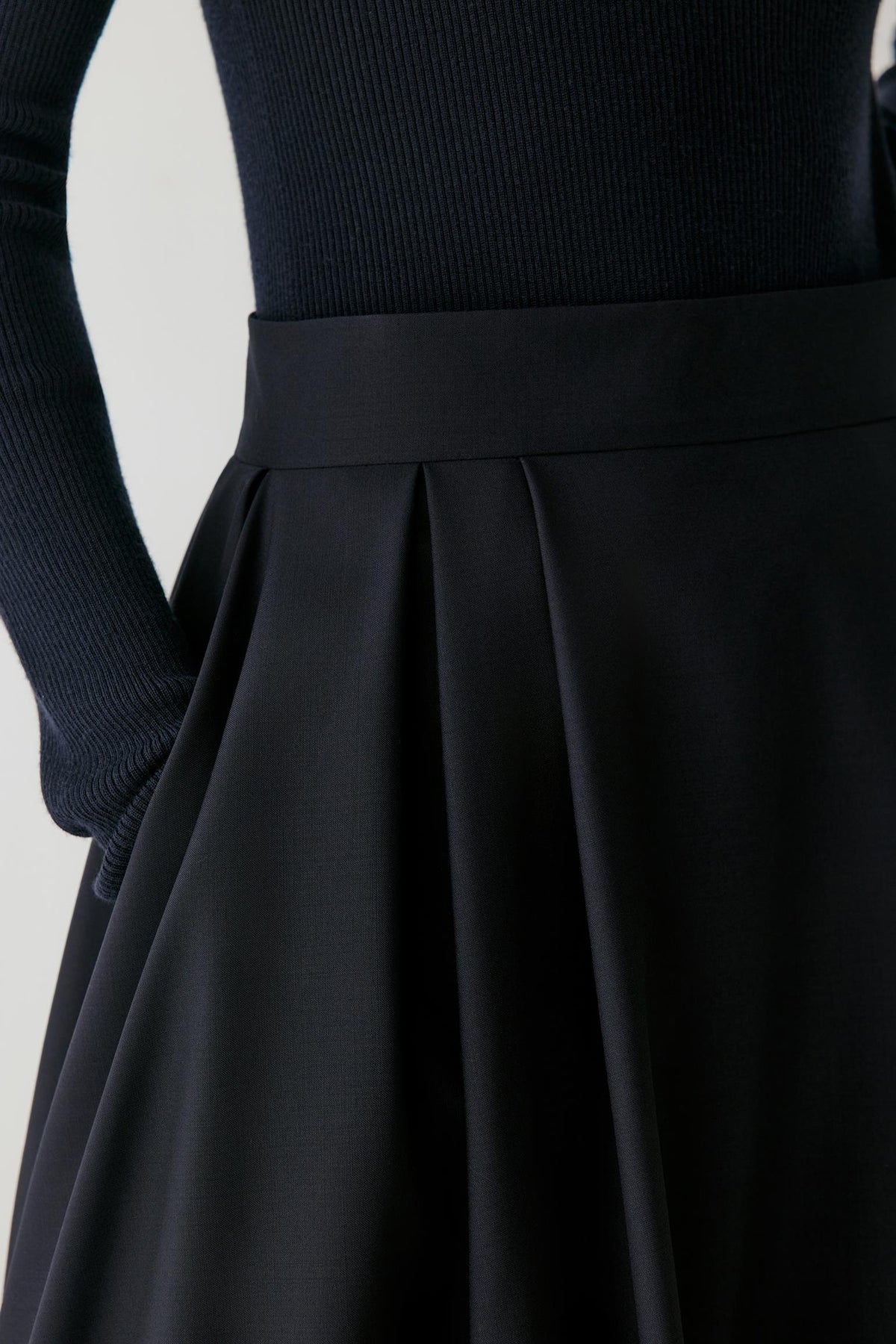 Close-up of a black pleated skirt with a wide waistband, paired with a ribbed black long-sleeve top, shown on a mannequin against a light gray background.