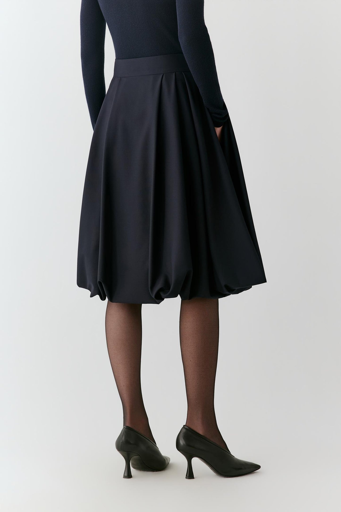 A person shown from the back wearing a dark navy pleated A-line skirt with a matching waistband, paired with black tights and black pointed-toe heels, against a light gray studio background.