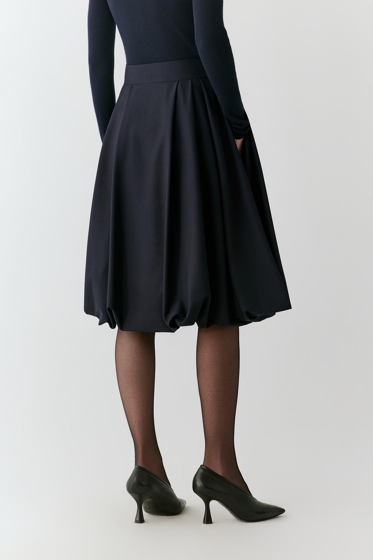 A person shown from the back wearing a dark navy pleated A-line skirt with a matching waistband, paired with black tights and black pointed-toe heels, against a light gray studio background.