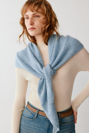 A woman wearing a light blue knit wrap or shawl tied around her shoulders over a cream ribbed sweater and blue jeans, photographed against a plain light background.