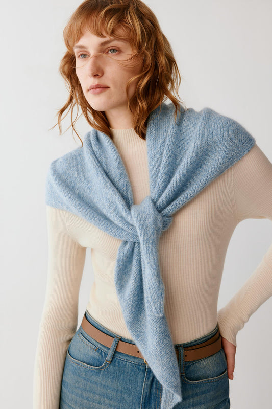 A woman wearing a light blue knit wrap or shawl tied around her shoulders over a cream ribbed sweater and blue jeans, photographed against a plain light background.