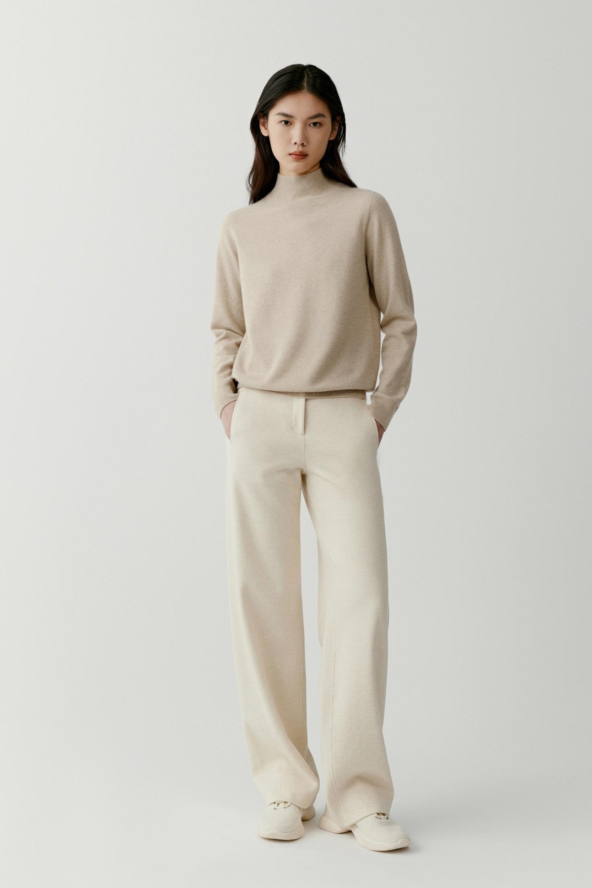 Front view of a woman wearing a beige cashmere pullover, showing the mock neck, dropped shoulders and relaxed silhouette.