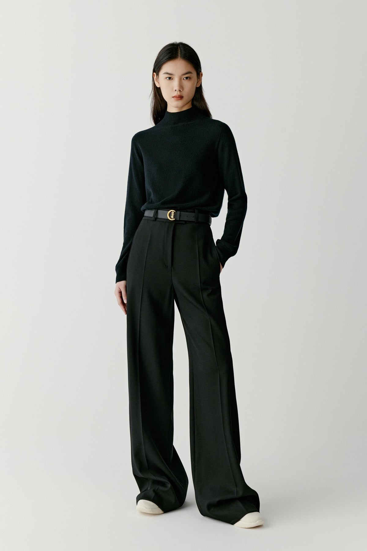 A model stands against a light gray studio background wearing a black mock-neck long-sleeve top, black wide-leg trousers, and a black belt with a gold buckle.