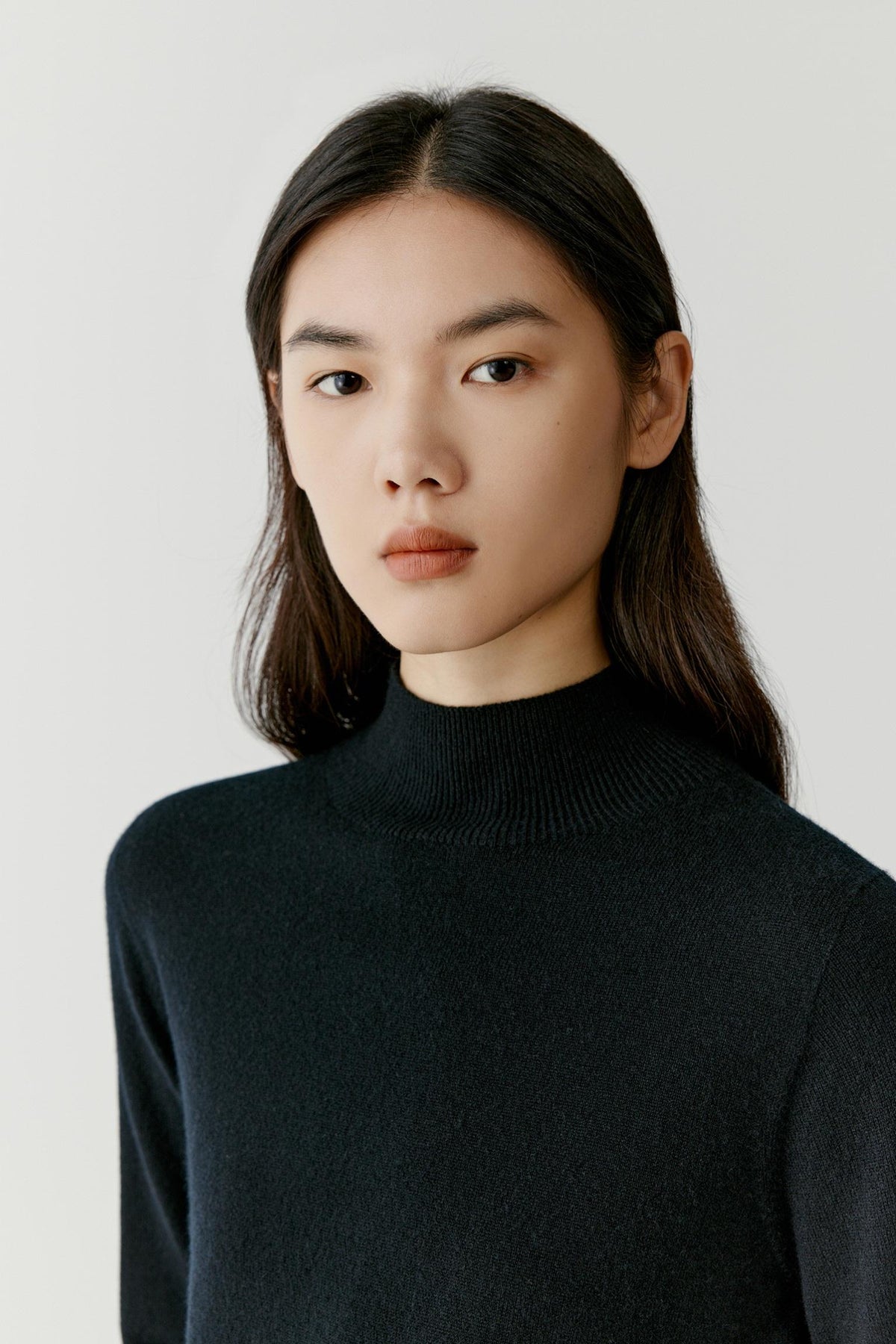 A woman with dark hair wearing a black ribbed mock neck sweater, photographed against a light gray studio background.