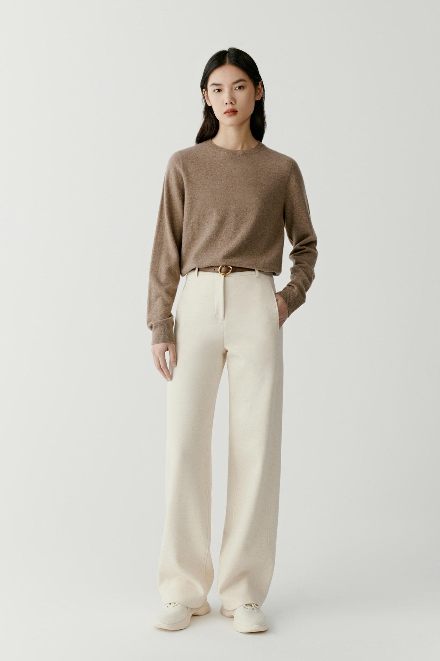 A model stands against a light gray studio background wearing a taupe-brown long-sleeve knit sweater, off-white wide-leg trousers, a thin brown belt with a gold buckle, and off-white slip-on shoes.