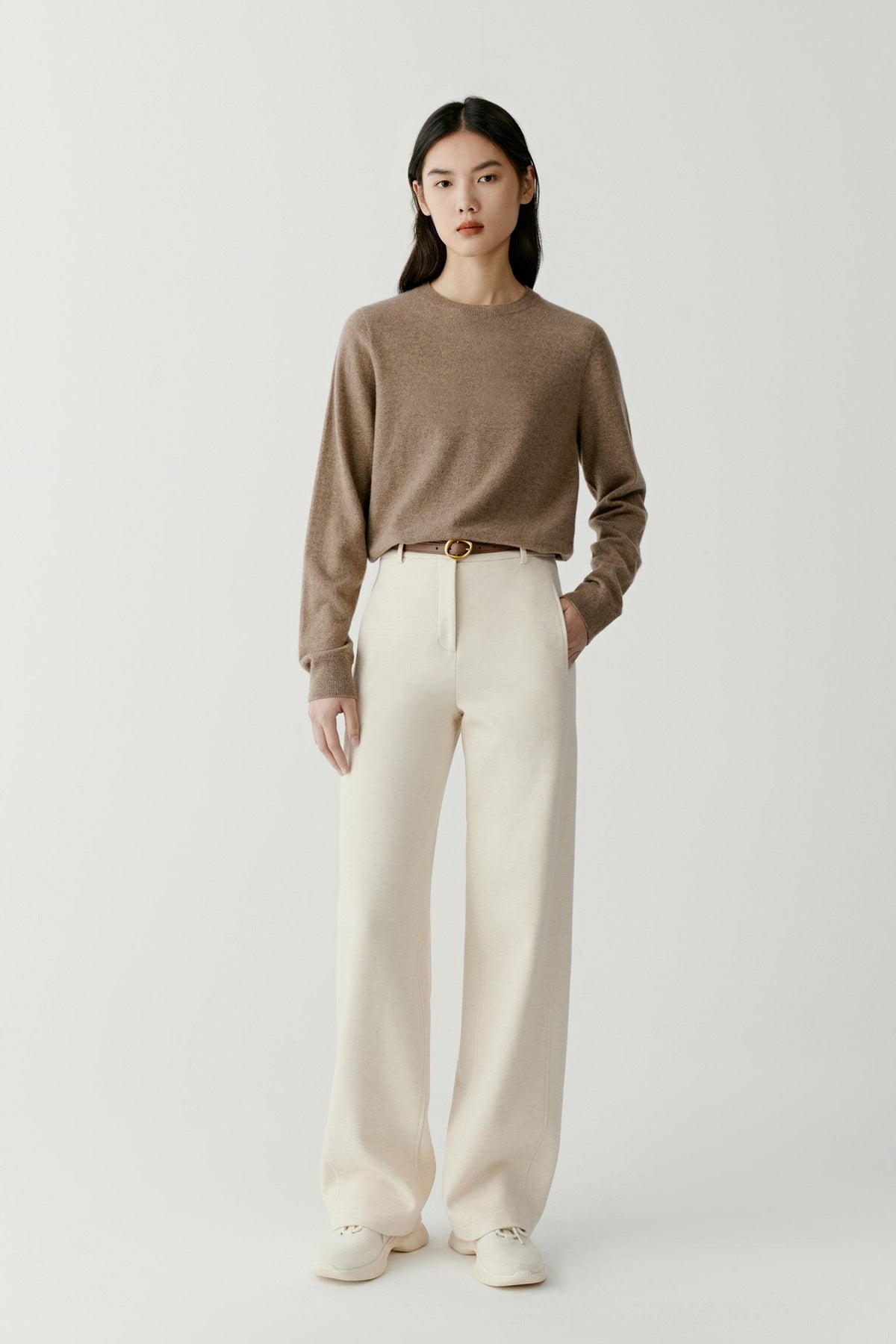 A model stands against a light gray studio background wearing a taupe-brown long-sleeve knit sweater, off-white wide-leg trousers, a thin brown belt with a gold buckle, and off-white slip-on shoes.