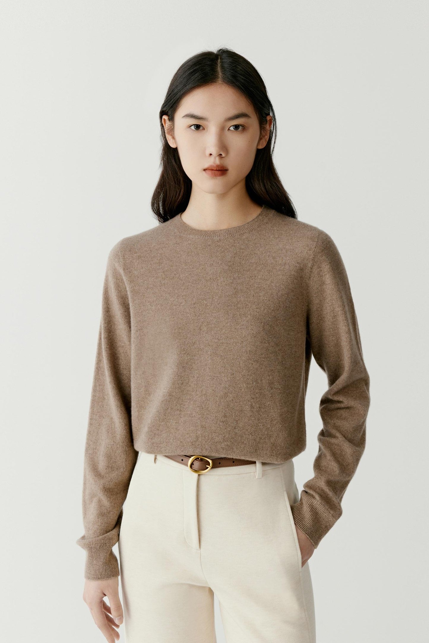 A model wearing a cropped, long-sleeve knit sweater in a warm taupe-brown color, paired with cream trousers and a thin brown belt with a gold buckle, against a light gray studio background.