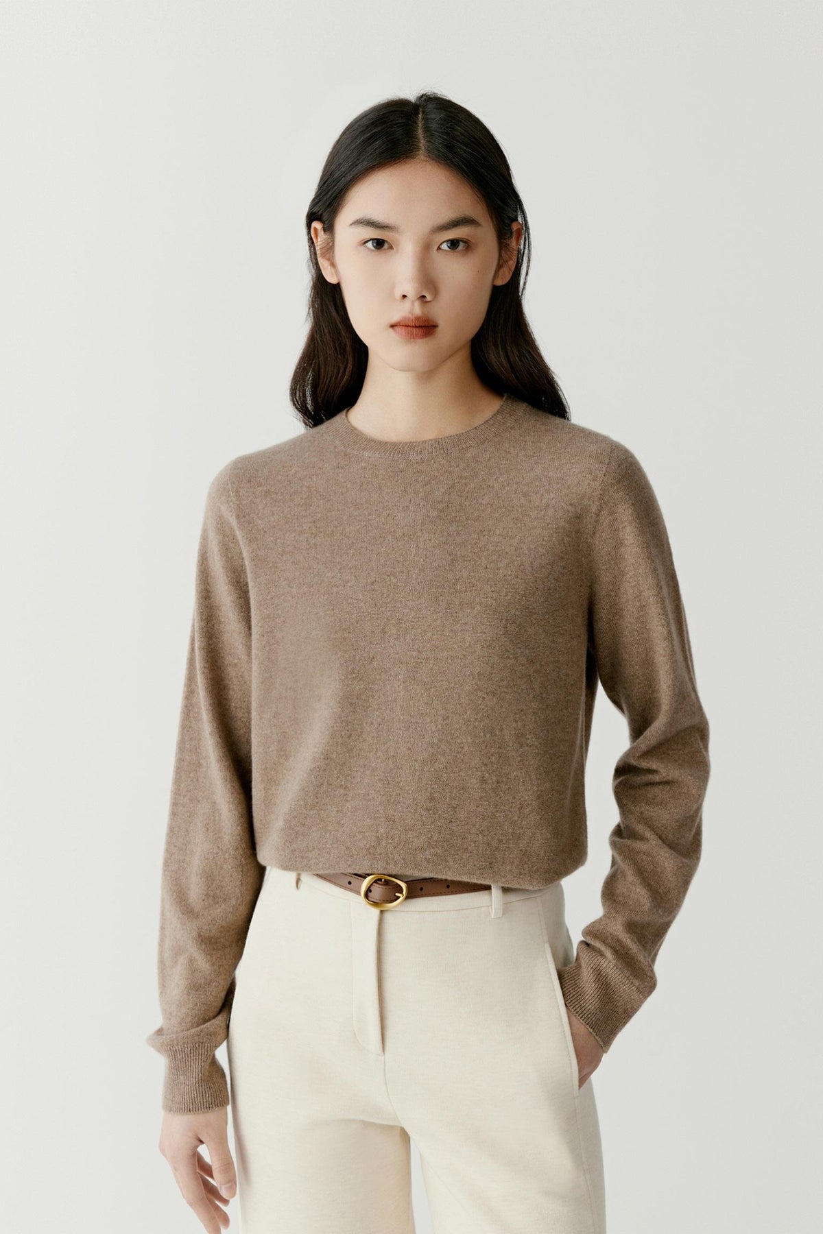 A model wearing a cropped, long-sleeve knit sweater in a warm taupe-brown color, paired with cream trousers and a thin brown belt with a gold buckle, against a light gray studio background.