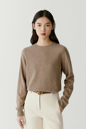 A model wearing a cropped, long-sleeve knit sweater in a warm taupe-brown color, paired with cream trousers and a thin brown belt with a gold buckle, against a light gray studio background.