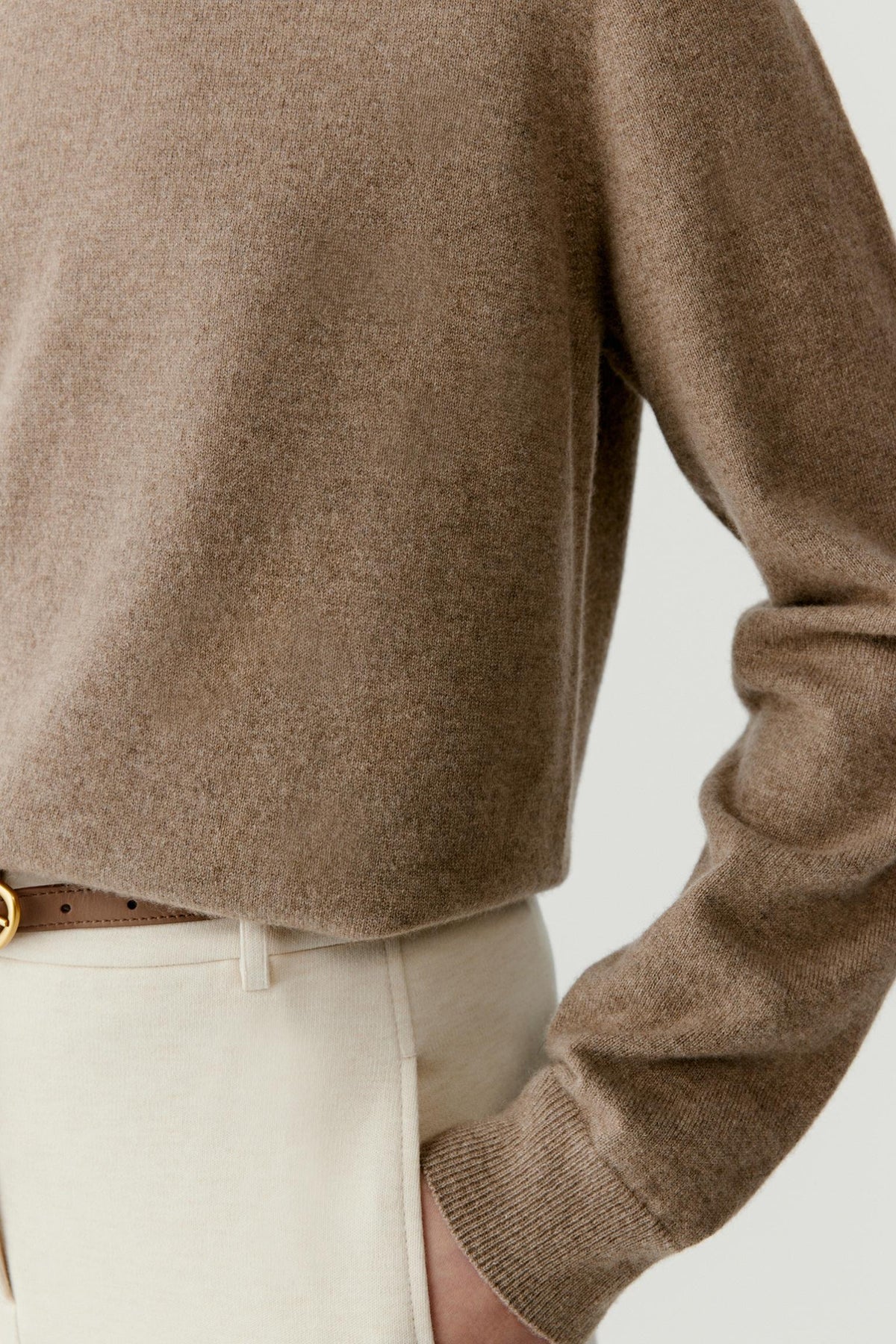 Close-up of a person wearing a taupe knit sweater with cream trousers and a brown belt, cropped to show the torso and arm against a plain light background.