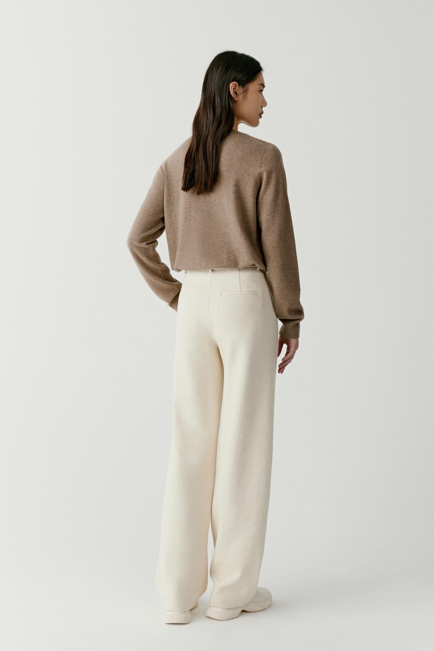 A woman is shown from the back wearing a cropped taupe knit sweater and cream wide-leg trousers, standing against a plain light gray studio background.