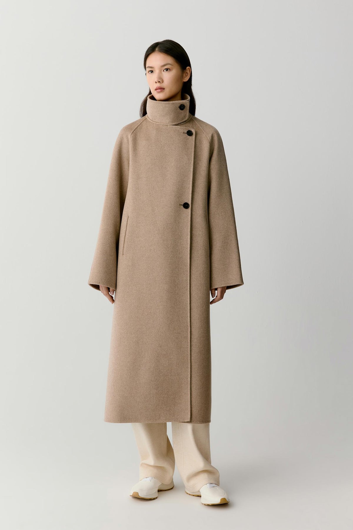 Front view of a woman wearing a natural beige double-face cashmere coat, showing the long silhouette, high stand collar and asymmetric button fastening.