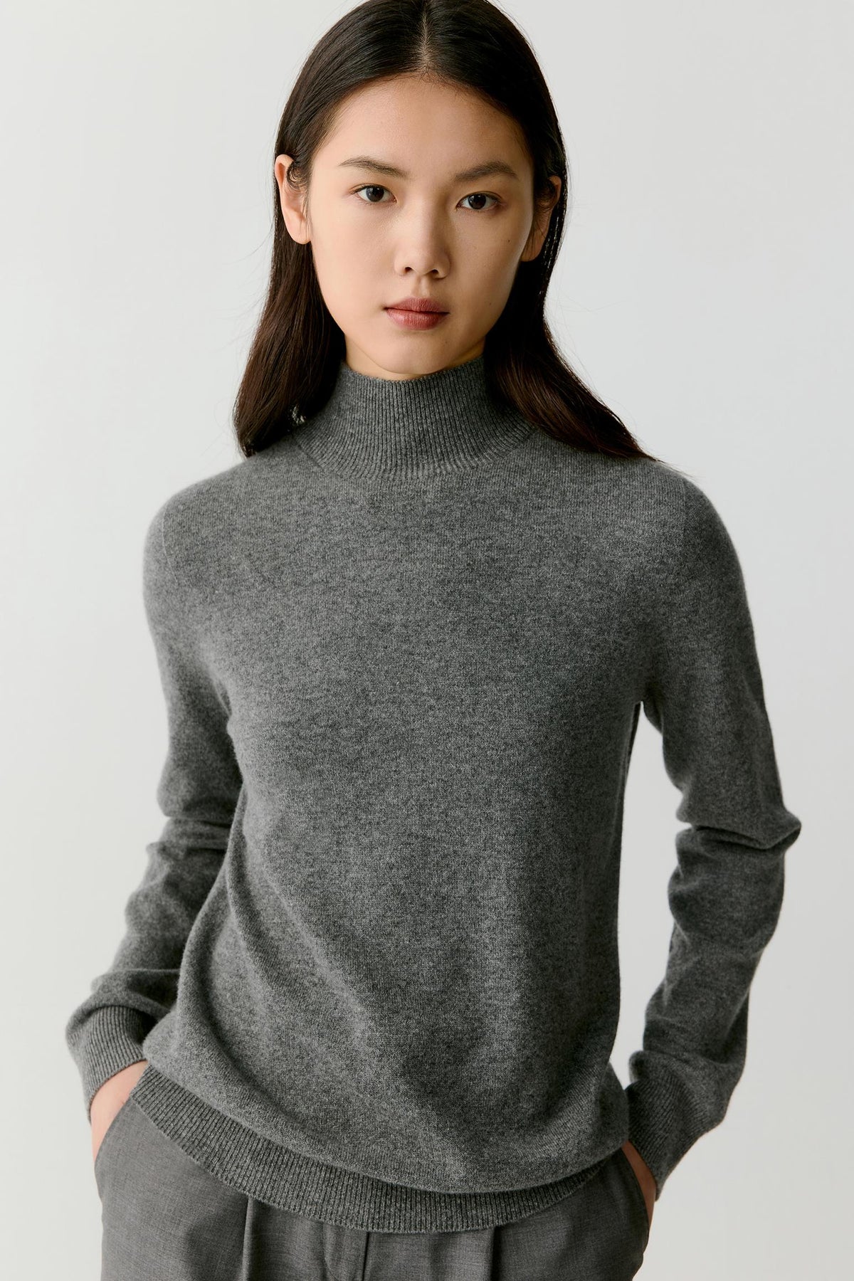 Front view of a woman wearing a smoky grey cashmere pullover, showing the mock neck, dropped shoulders and relaxed silhouette.
