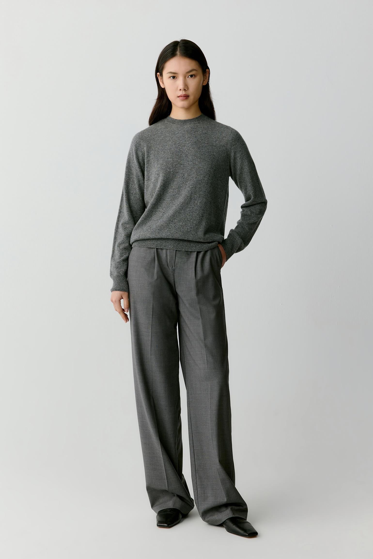 A model stands against a light gray studio background wearing a dark gray knit crewneck sweater and matching dark gray wide-leg trousers.
