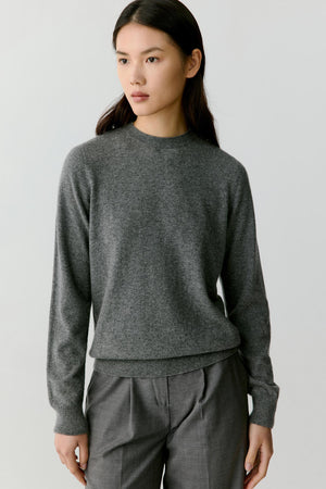 A woman wearing a dark gray knit crewneck sweater and matching gray trousers, photographed against a plain light gray studio background.