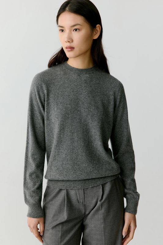 A woman wearing a dark gray knit crewneck sweater and matching gray trousers, photographed against a plain light gray studio background.