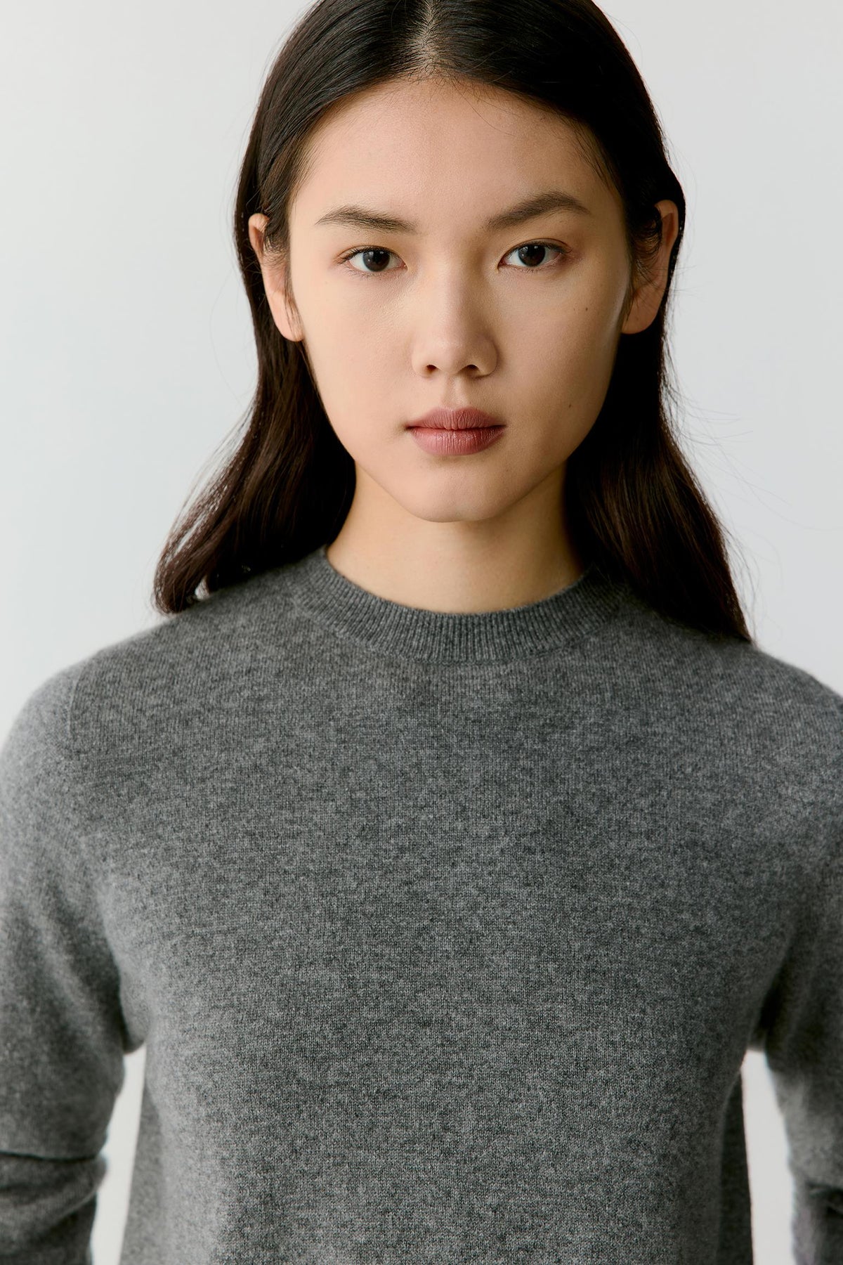 A woman wearing a gray knit crewneck sweater, shown from the chest up against a plain light gray background.