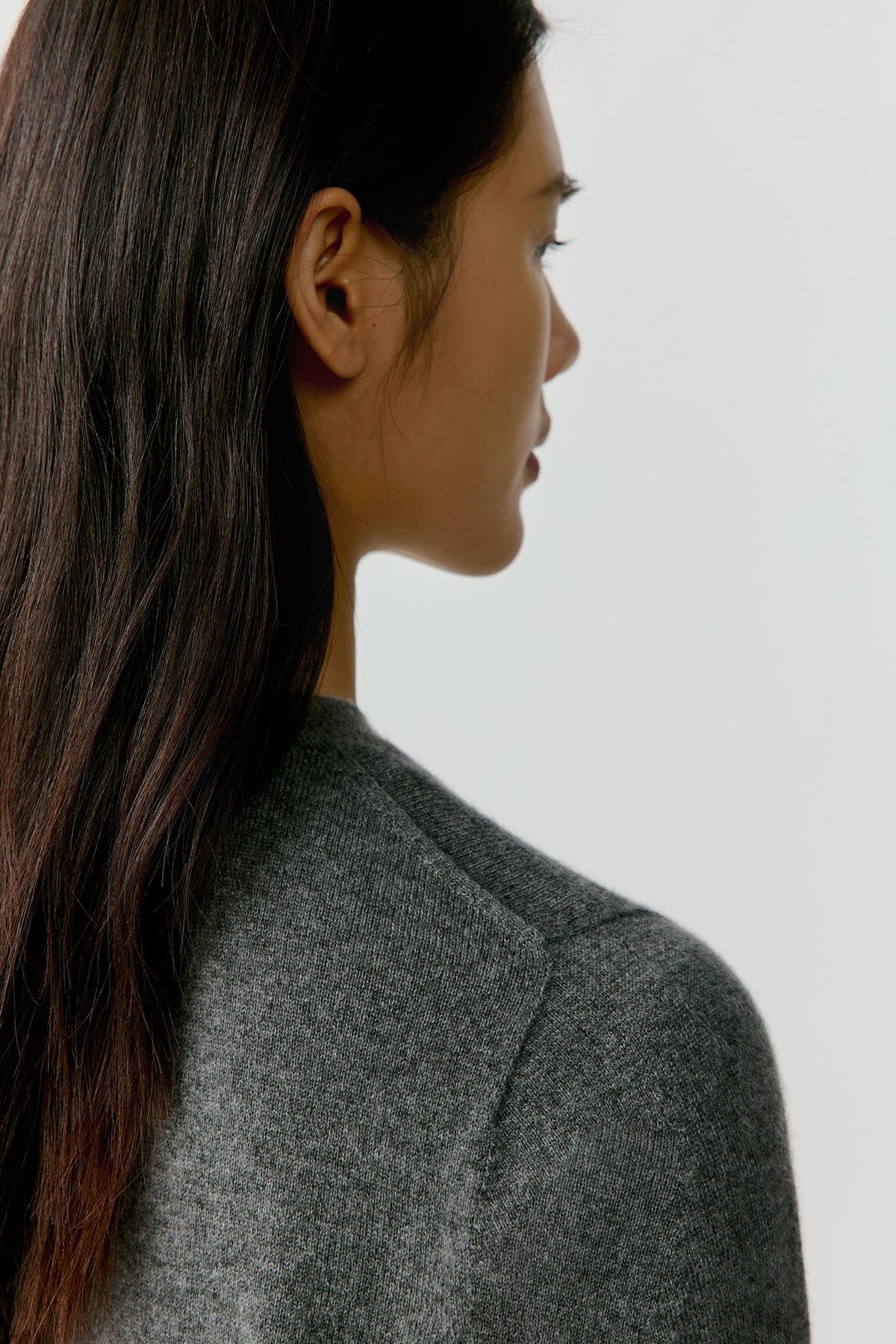 A close-up side profile of a woman with long dark hair wearing a textured gray knit top against a plain light gray background.