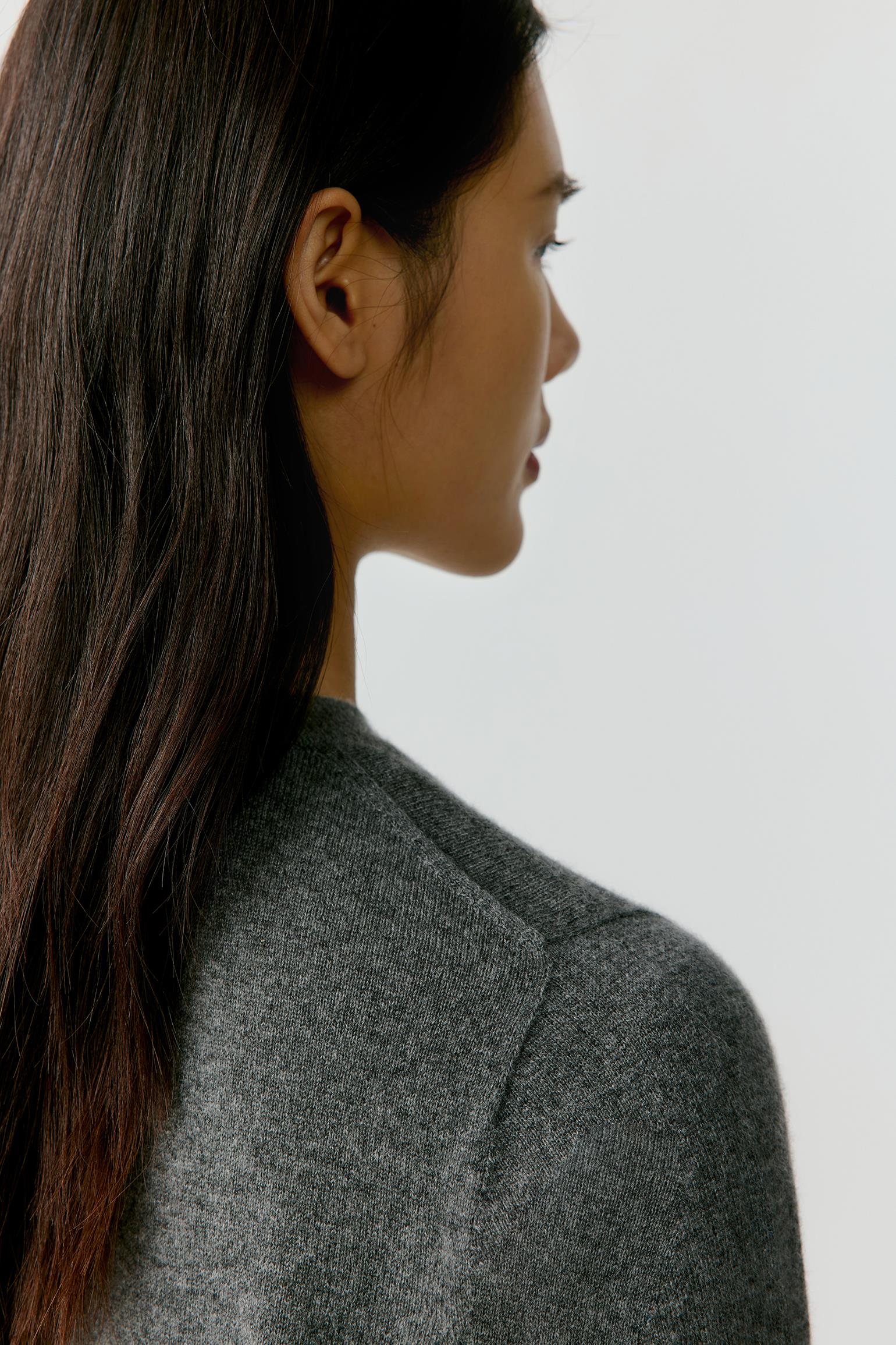 A close-up side profile of a woman with long dark hair wearing a textured gray knit top against a plain light gray background.