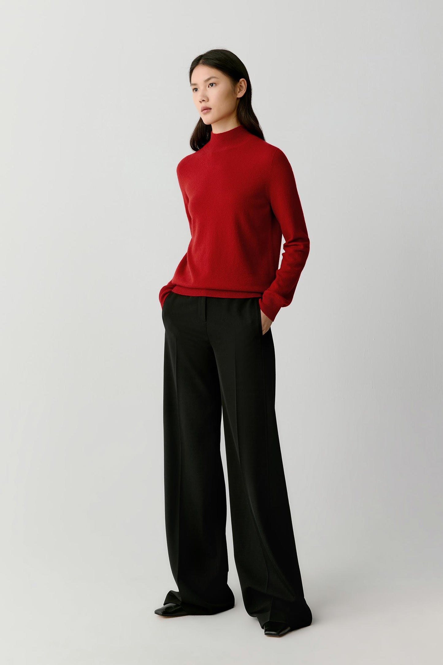 A woman modeling a red mock-neck knit sweater paired with black wide-leg trousers, standing against a light gray studio background.