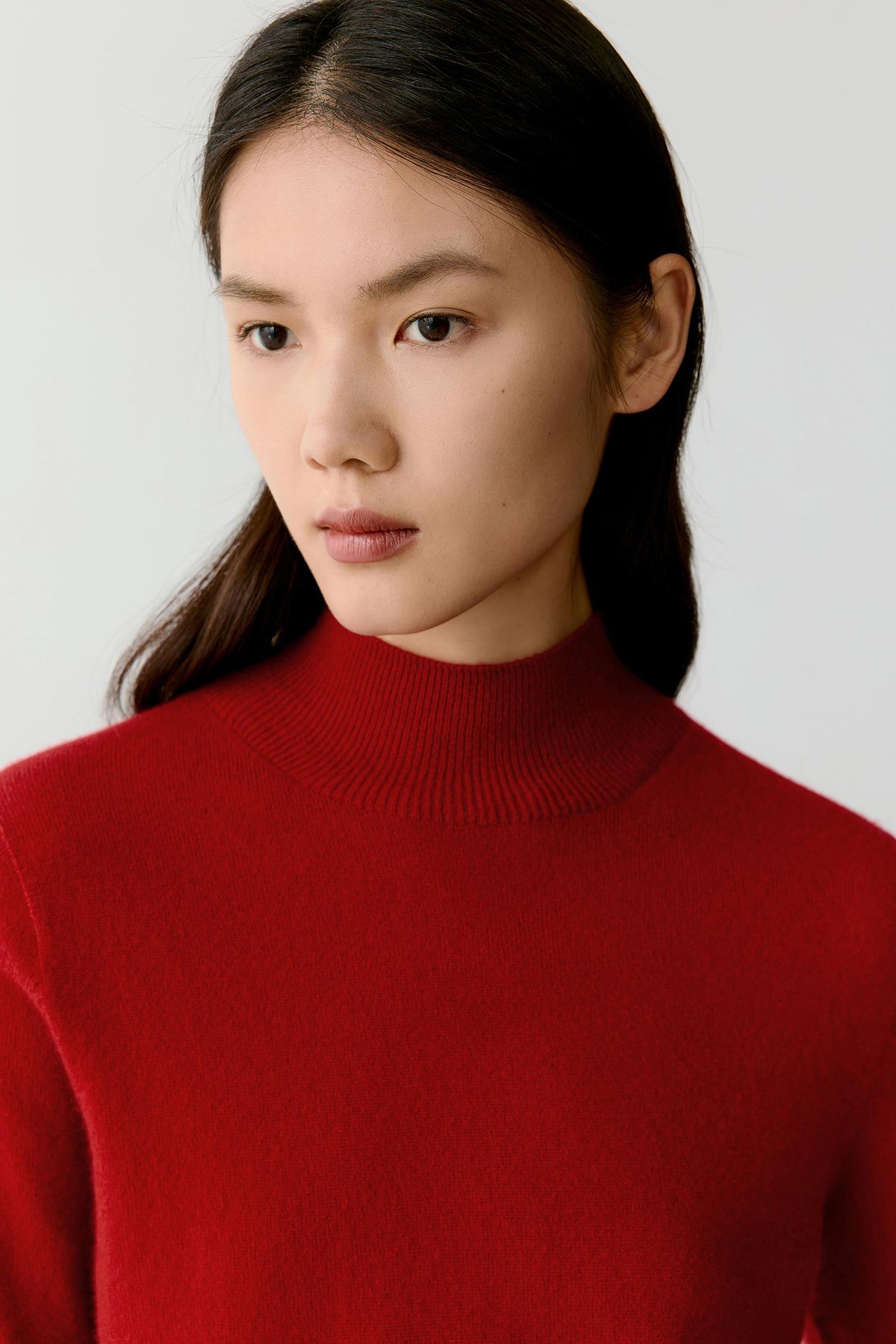 A close-up portrait of a woman wearing a bright red ribbed mock neck sweater against a light gray background.