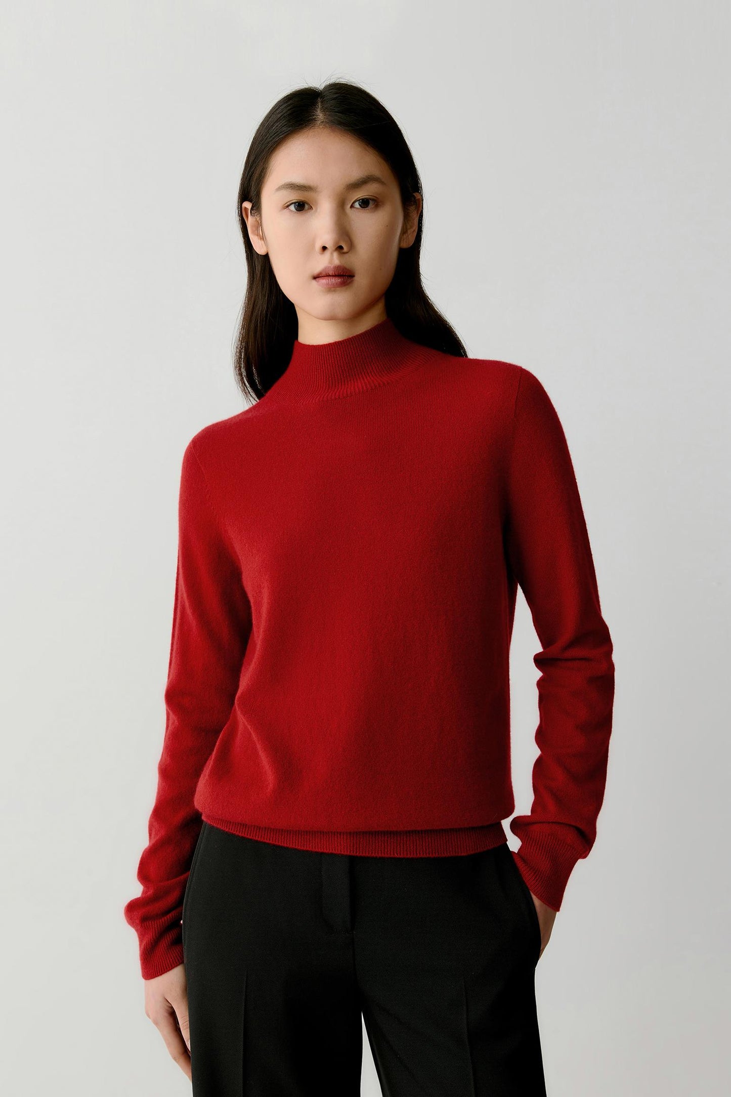 A woman modeling a red mock-neck knit sweater with black trousers, standing against a light gray studio background.