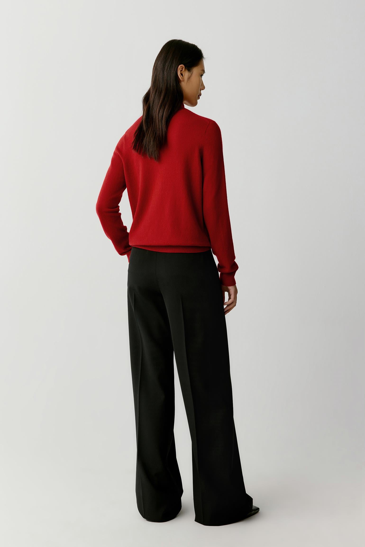 A woman shown from the back wearing a bright red long-sleeve knit sweater and black wide-leg trousers, standing against a plain light gray studio background.