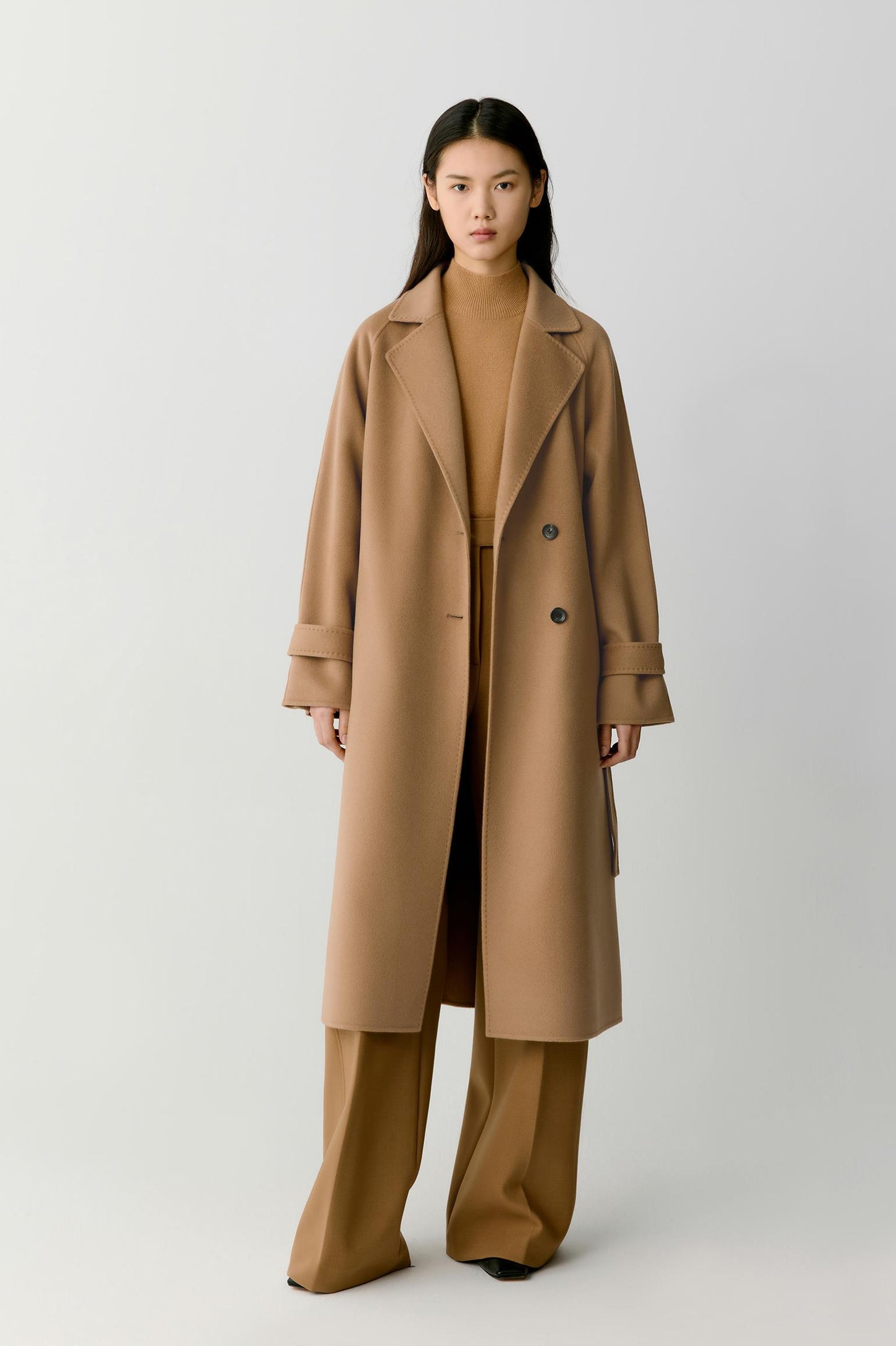 A model stands against a light gray studio background wearing a long camel-brown double-breasted coat over a matching tan top and wide-leg trousers.