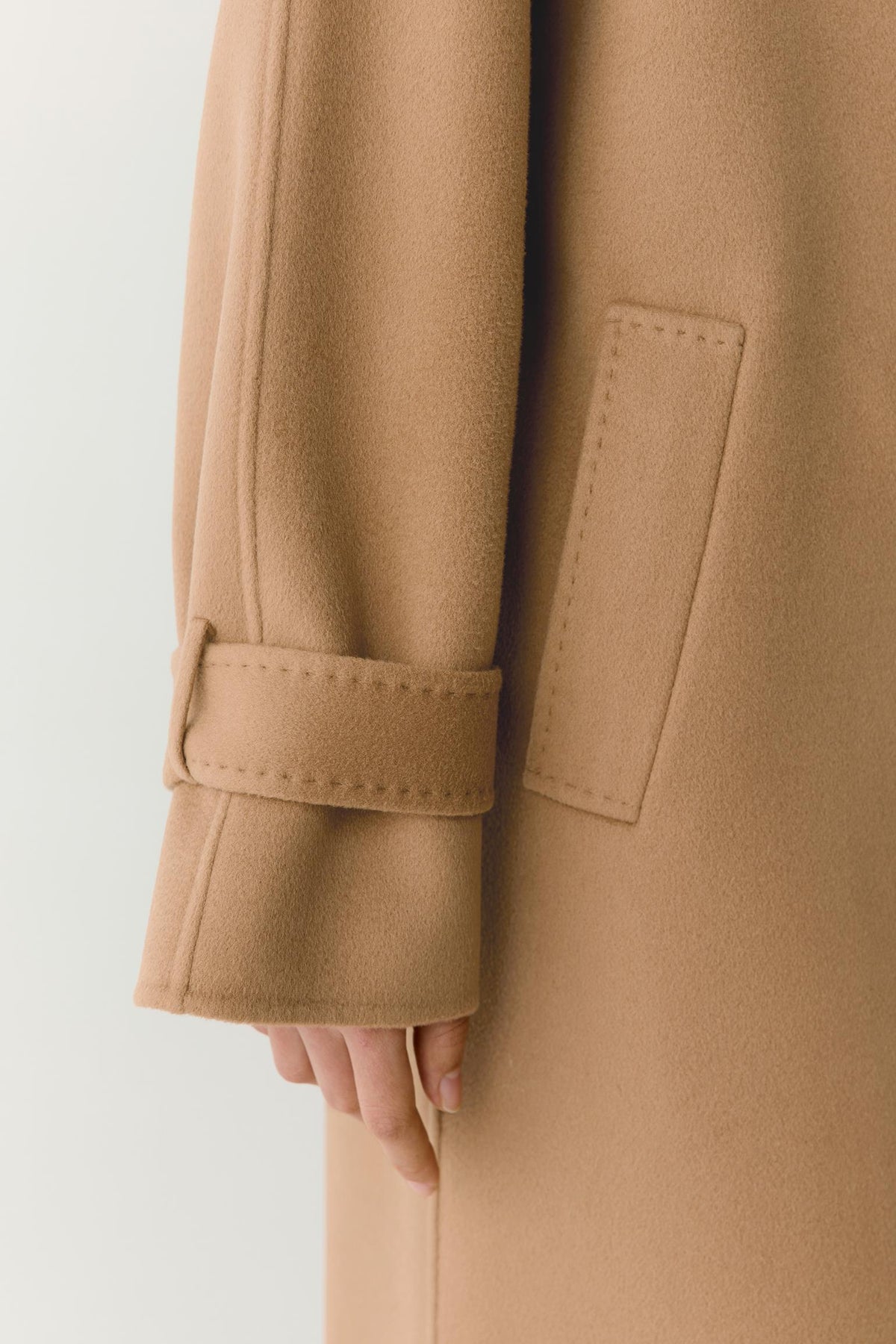 Close-up of a camel-colored wool coat sleeve with a matching cuff strap and a side welt pocket, shown on a model against a light gray background.