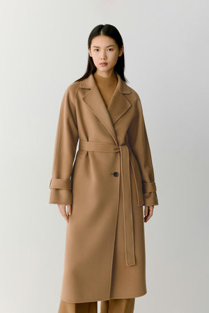 A woman modeling a long camel-brown belted coat over a matching brown top and trousers, standing against a plain light gray studio background.