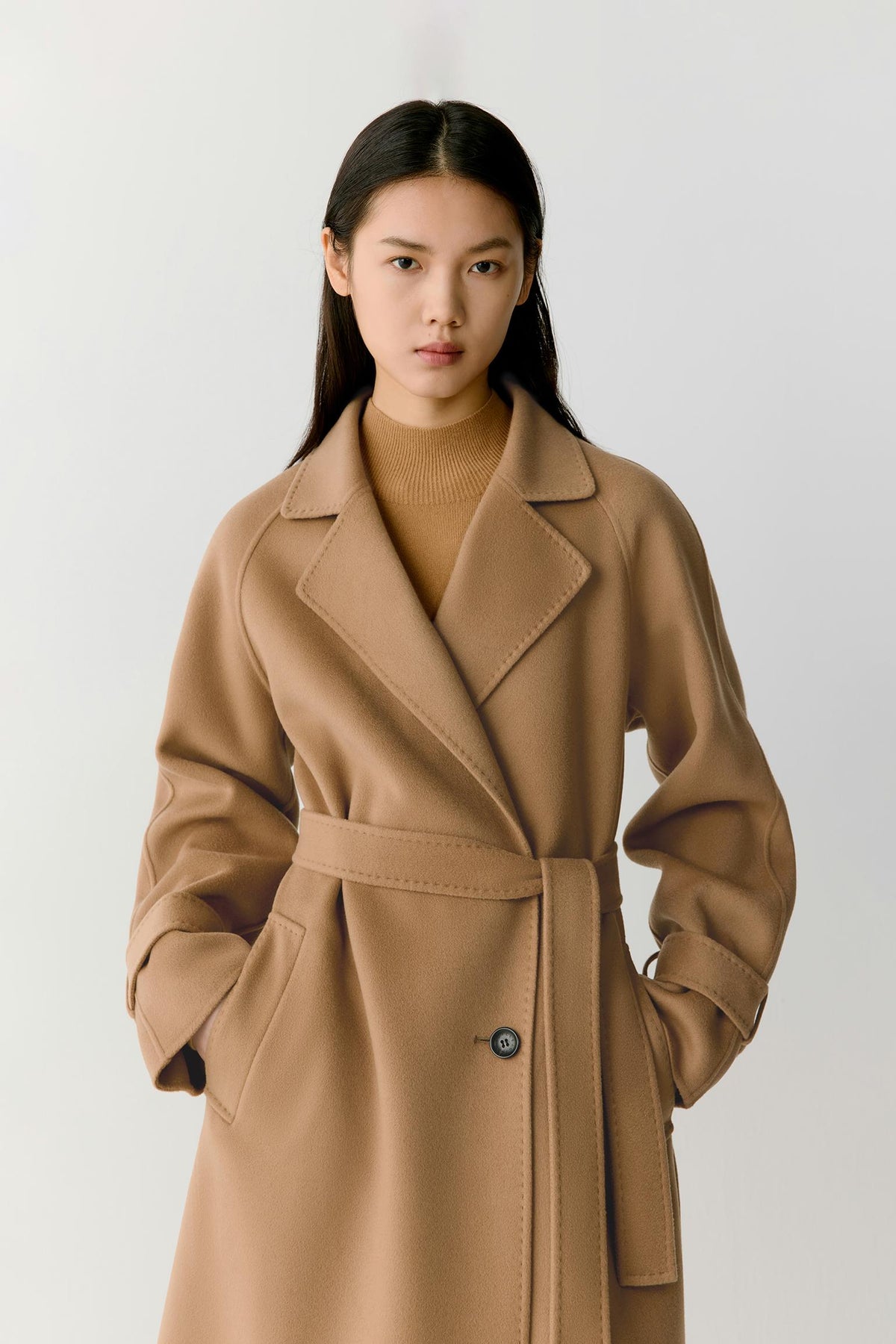 A woman wearing a camel-colored belted coat over a matching ribbed knit top, photographed against a plain light gray studio background.