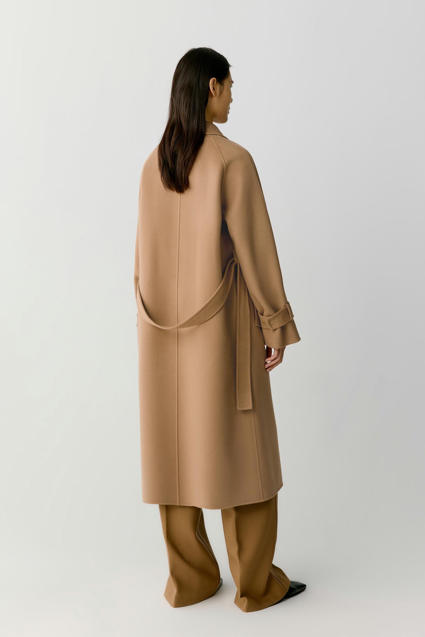 A model shown from the back wearing a long camel-colored wrap coat and matching wide-leg trousers against a plain light gray studio background.