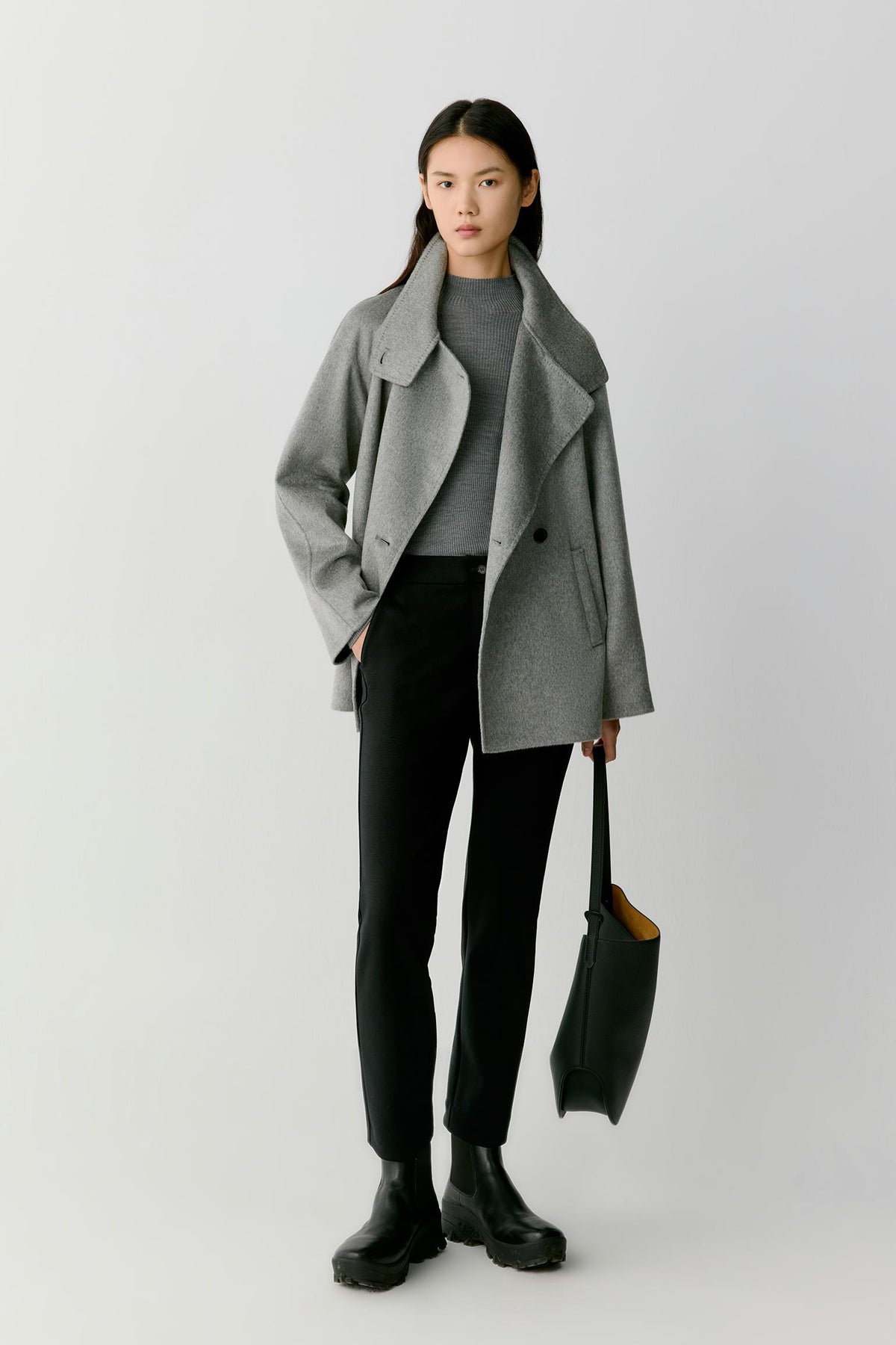 Front view of a woman wearing a grey melange double-face cashmere short coat, showing the high stand collar, asymmetric button fastening and relaxed silhouette.