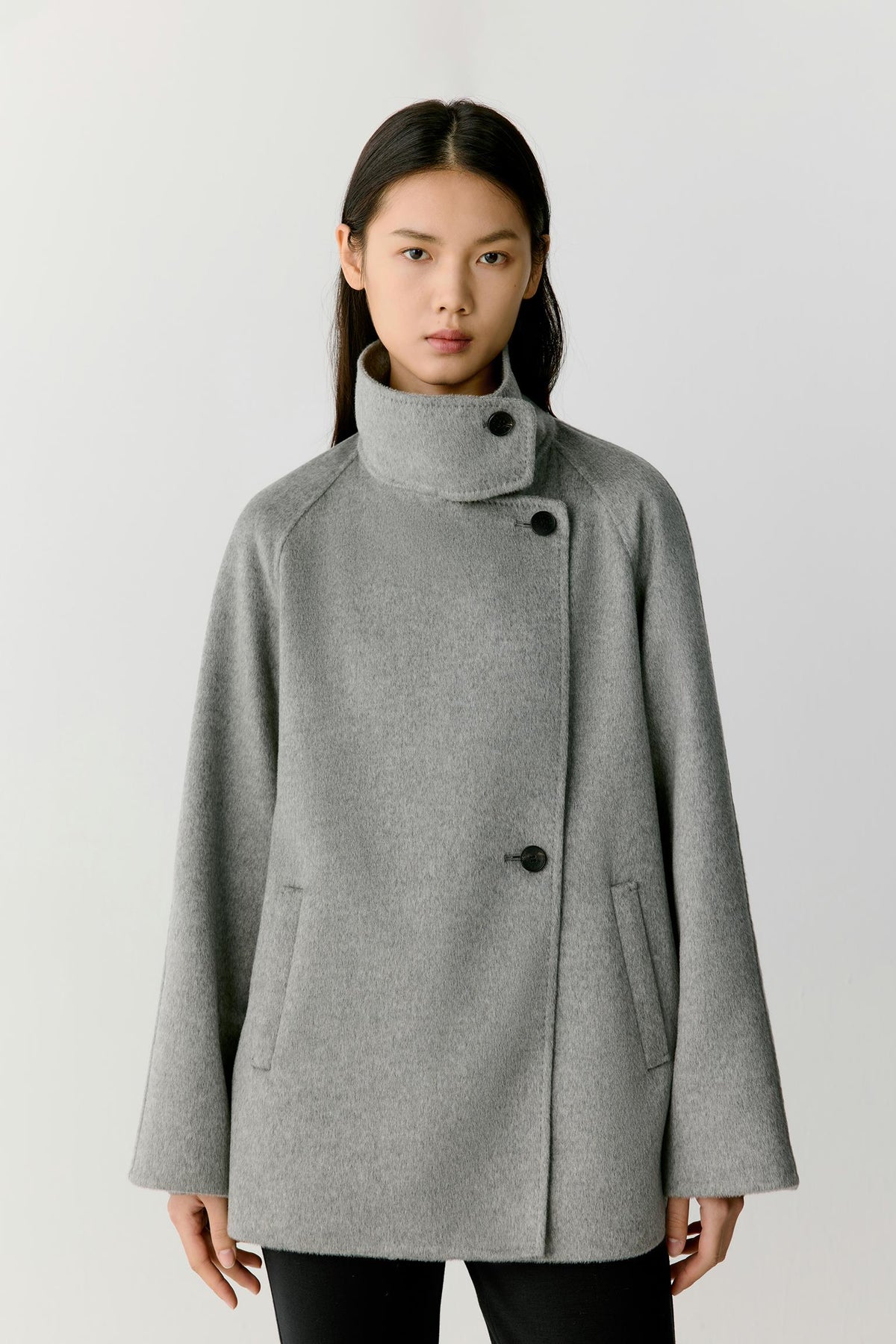 Front view of a woman wearing a grey melange double-face cashmere short coat, showing the high stand collar, asymmetric button fastening and relaxed silhouette.