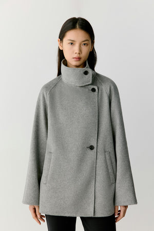 Front view of a woman wearing a grey melange double-face cashmere short coat, showing the high stand collar, asymmetric button fastening and relaxed silhouette.