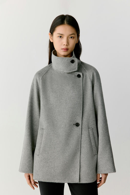 Front view of a woman wearing a grey melange double-face cashmere short coat, showing the high stand collar, asymmetric button fastening and relaxed silhouette.