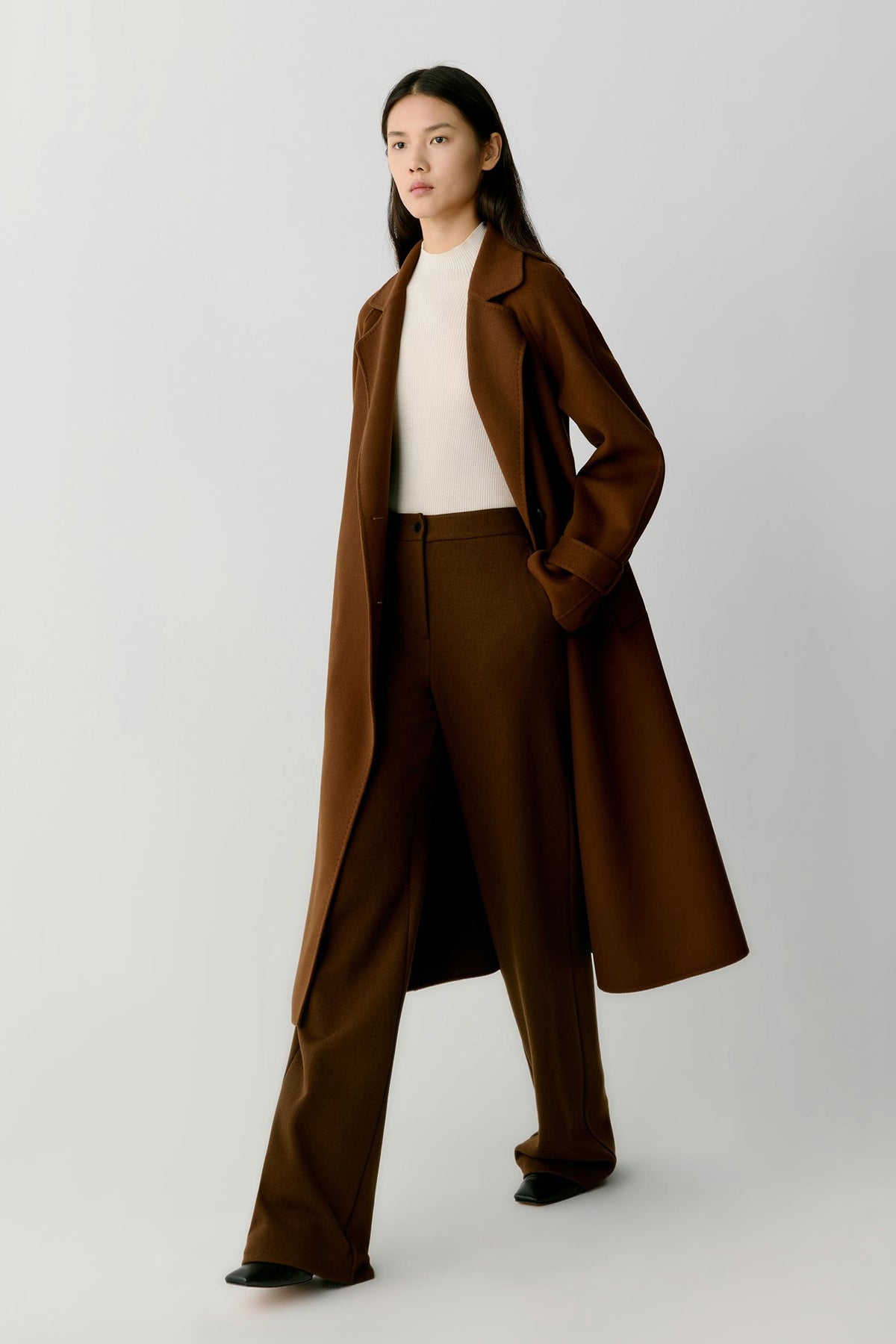 A model wearing a long brown coat, matching brown trousers, a cream ribbed top, and black shoes, standing against a light gray studio background.