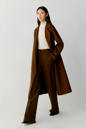 A model wearing a long brown coat, matching brown trousers, a cream ribbed top, and black shoes, standing against a light gray studio background.