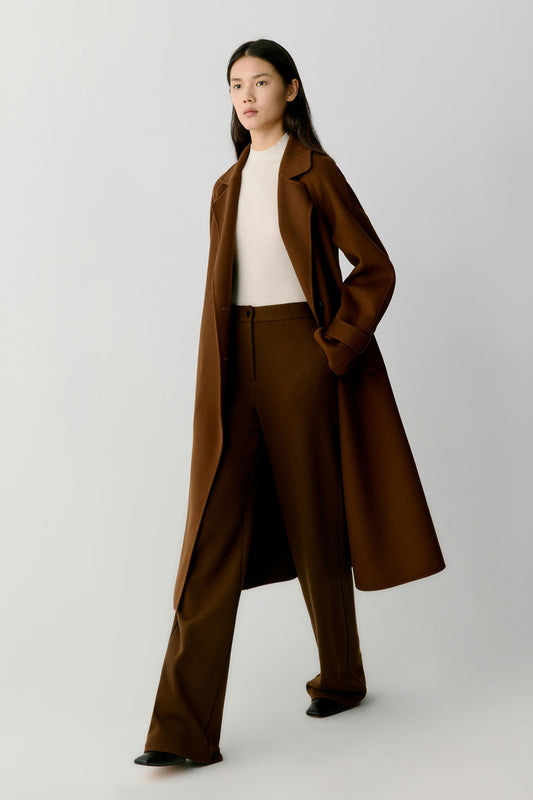 A model wearing a long brown coat, matching brown trousers, a cream ribbed top, and black shoes, standing against a light gray studio background.