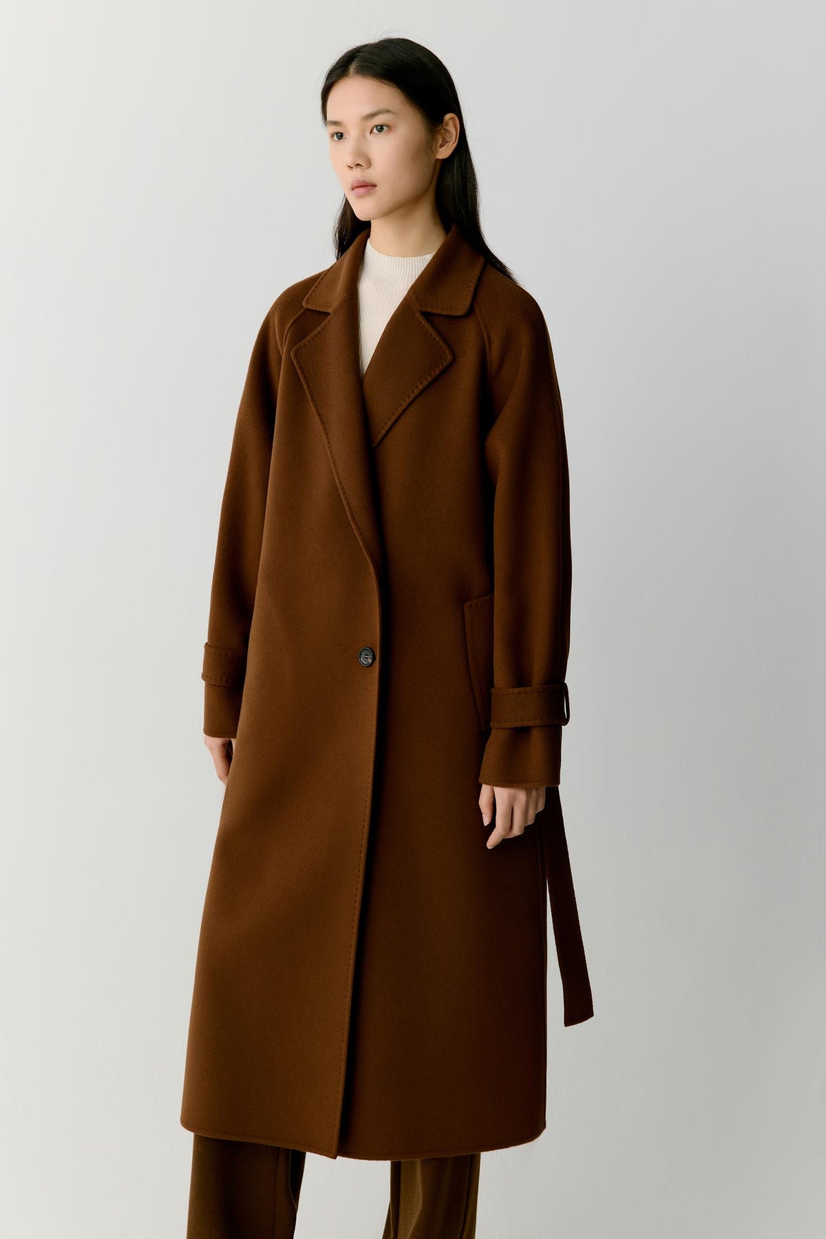 A woman modeling a long brown wool-blend coat with a single button closure, notched lapels, and a self-tie belt, paired with matching brown trousers and a cream top, against a light gray studio background.