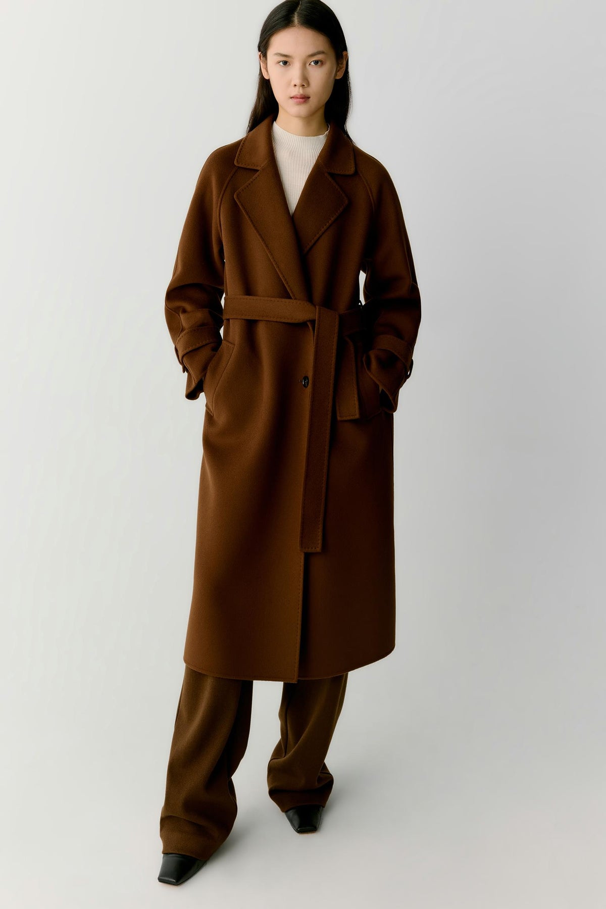 A model wearing a long brown belted coat over a cream top and matching brown trousers, standing against a light gray studio background.