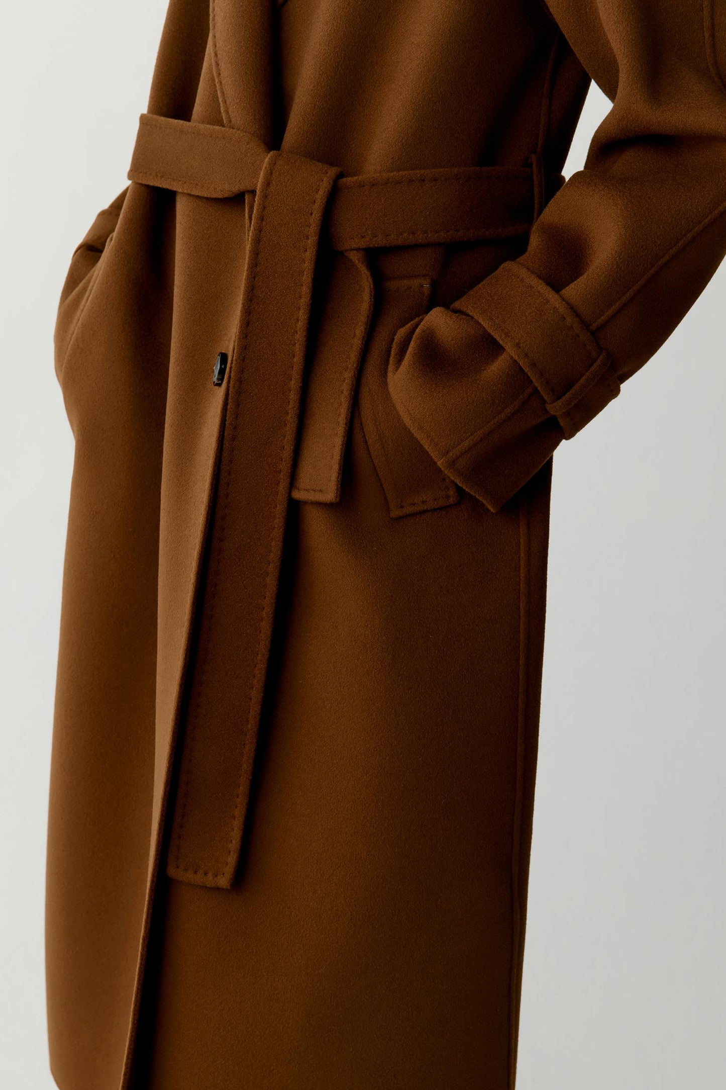 Close-up of a brown wool coat with a matching belt, featuring a sleeve with a decorative tab and a pocket, set against a plain light gray background.
