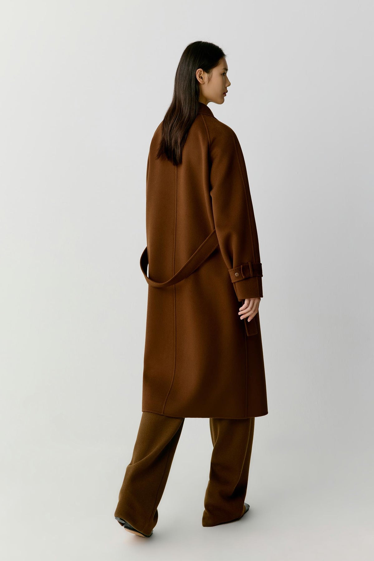 A woman is shown from the back wearing a long brown coat and matching wide-leg trousers against a plain light gray studio background.