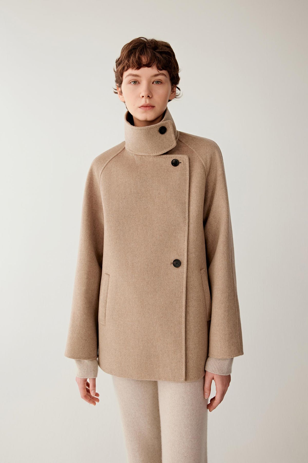 Three-quarter view highlighting the short cut, raglan sleeves and structured drape of the natural beige double-face cashmere coat.