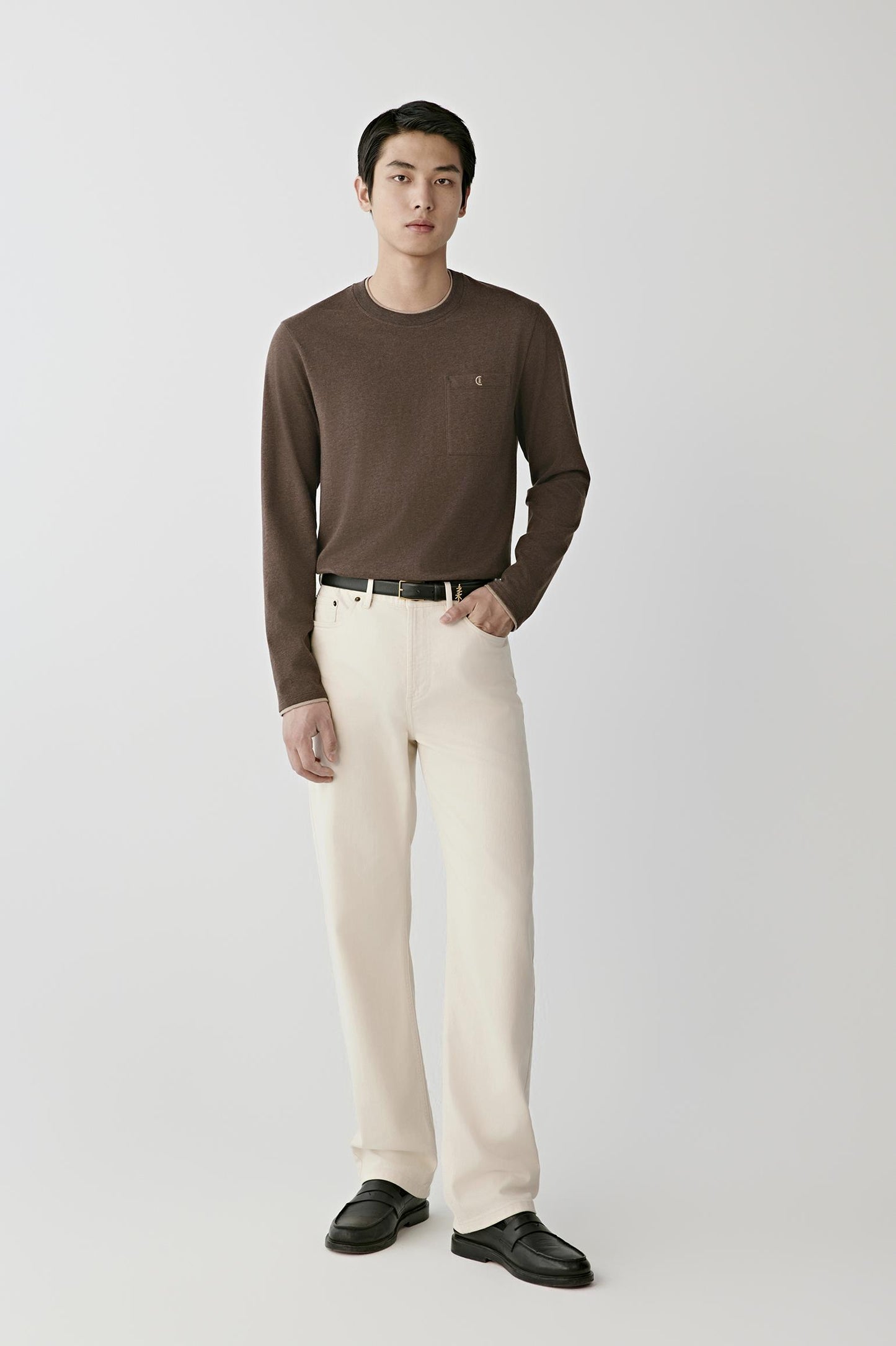 A man modeling a dark brown long-sleeve crewneck sweater paired with cream-colored trousers, a black belt, and black leather shoes, standing against a light gray studio background.