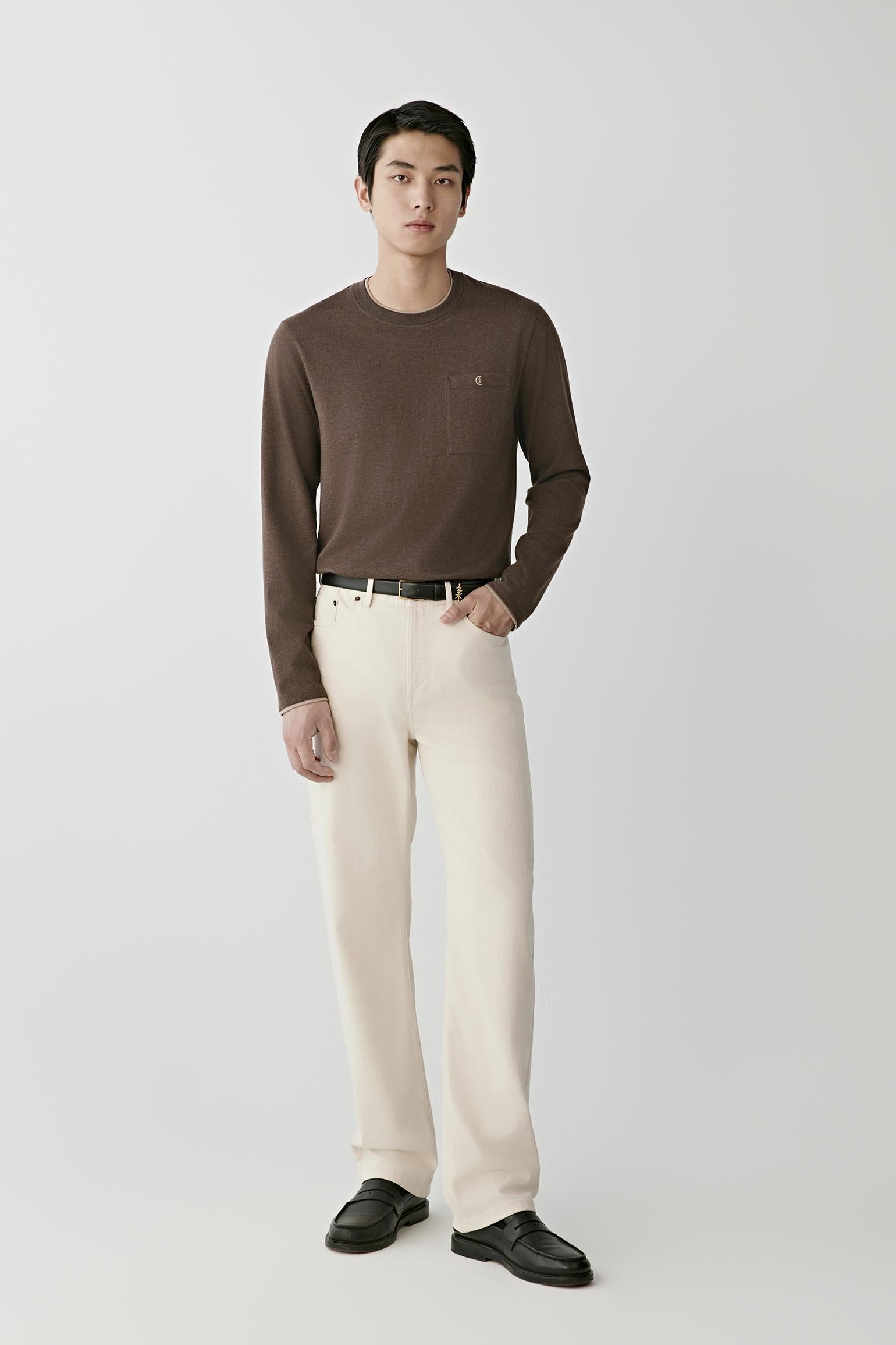 A man modeling a dark brown long-sleeve crewneck sweater paired with cream-colored trousers, a black belt, and black leather shoes, standing against a light gray studio background.